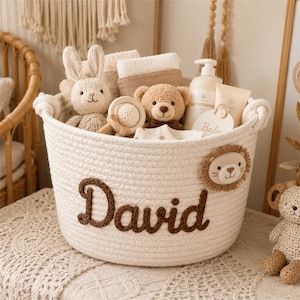 Puede incluir: Una cesta de cuerda trenzada blanca con el nombre David en letras cursivas marrones y un parche de cara de león. La cesta contiene un conejo de punto, un osito de peluche, un sonajero de madera, botellas de cuidado infantil y una etiqueta Baby.