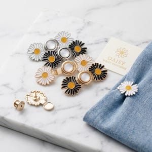 May include: A set of daisy-shaped waist cinchers featuring white and black enamel petals with yellow centers and metallic backings. The set includes a small card labeled DAISY WAIST CINCHERS and one cincher attached to a piece of blue denim fabric.
