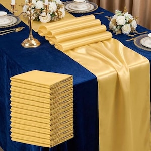May include: A set of gold satin table runners and matching napkins arranged on a deep blue velvet tablecloth. The runners feature a smooth, reflective finish, while the folded napkins are stacked in a neat pile, creating a contrast of textures.