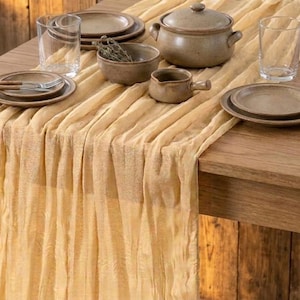 May include: A light yellow, crinkled fabric table runner draped over a wooden table, set with ceramic plates, a lidded pot, a small bowl, two clear glass tumblers, and a metal fork resting on a plate, creating a coordinated dining arrangement.