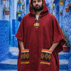 May include: A burgundy hooded poncho with patterned accents on the hood, pockets, and hem. The poncho features a button closure at the neck and is worn in a blue-walled alleyway. The pockets have a geometric design with orange, yellow, and black details.