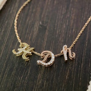 May include: A gold-colored necklace with three cursive letters: A, D, and h. Each letter is embellished with small, clear gemstones. The necklace chain is delicate, and the letters are connected. The background is a dark wood surface.
