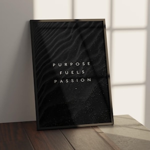 May include: A framed black and white art print with the words "PURPOSE FUELS PASSION" in white lettering. The background features a textured, dark pattern. The frame is black, and the print is leaning against a white wall.