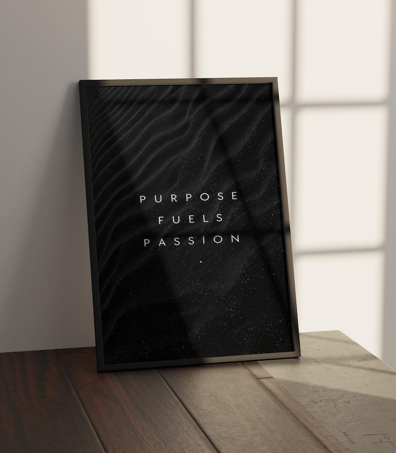 May include: A framed black and white art print with the words "PURPOSE FUELS PASSION" in white lettering. The background features a textured, dark pattern. The frame is black, and the print is leaning against a white wall.