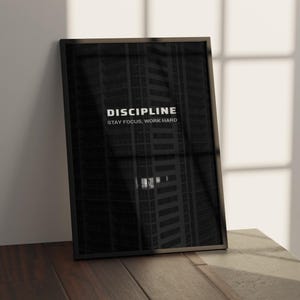 May include: A framed black and white print with the word "DISCIPLINE" at the top and "STAY FOCUS, WORK HARD" below it. The background is a dark image of a tall building. The frame is black and the print is leaning against a wall.