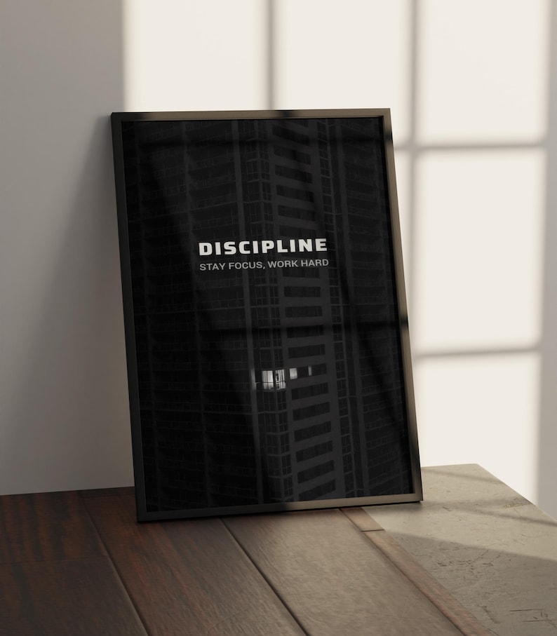 May include: A framed black and white print with the word "DISCIPLINE" at the top and "STAY FOCUS, WORK HARD" below it. The background is a dark image of a tall building. The frame is black and the print is leaning against a wall.