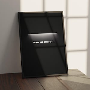 May include: A framed black poster with the words "now or never" in white. The poster is leaning against a white wall and is on a wooden surface. A horizontal white light illuminates the center of the poster.