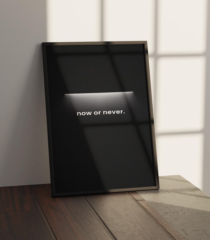 May include: A framed black poster with the words "now or never" in white. The poster is leaning against a white wall and is on a wooden surface. A horizontal white light illuminates the center of the poster.