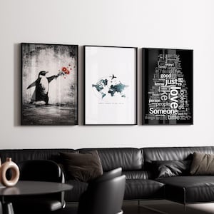 Op de afbeelding: Drie ingelijste kunstdrukken. De eerste toont een pinguïn met bloemen, de tweede een wereldkaart met een vliegtuig en de derde een woordwolk met woorden als "liefde" en "tijd". De lijsten zijn zwart.