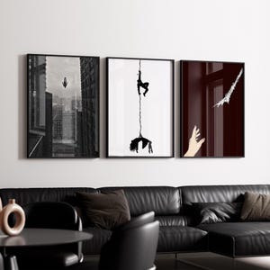 May include: Three framed art prints. The first is a black and white cityscape with a figure falling. The second features a figure suspended from a rope. The third has a hand reaching towards a white shape.