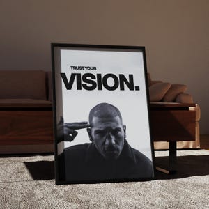 May include: Black and white framed poster with the text "TRUST YOUR VISION." The image features a man with a serious expression, with a hand gesturing a finger gun to his head. The poster is leaning against a wall.