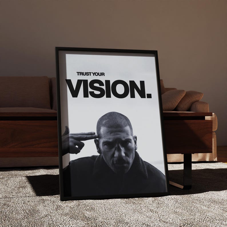 May include: Black and white framed poster with the text "TRUST YOUR VISION." The image features a man with a serious expression, with a hand gesturing a finger gun to his head. The poster is leaning against a wall.