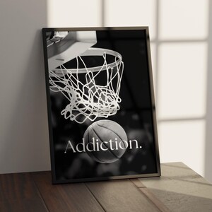 May include: Black and white framed print featuring a basketball going through a net. The word "Addiction" is printed in white below the ball. The print is leaning against a wall on a wooden surface.