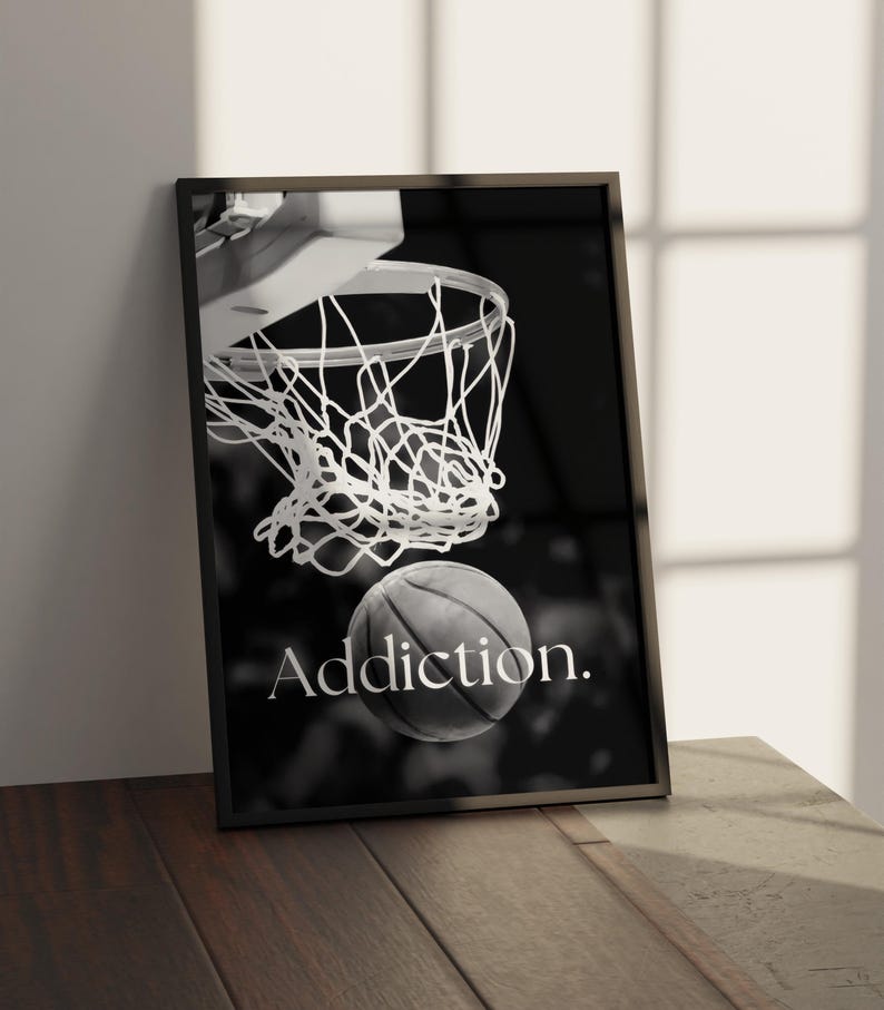 May include: Black and white framed print featuring a basketball going through a net. The word "Addiction" is printed in white below the ball. The print is leaning against a wall on a wooden surface.