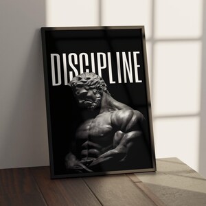 May include: Framed black and white print featuring a muscular statue of a man. The word "DISCIPLINE" is printed in large white letters at the top. The print is displayed on a wooden surface.