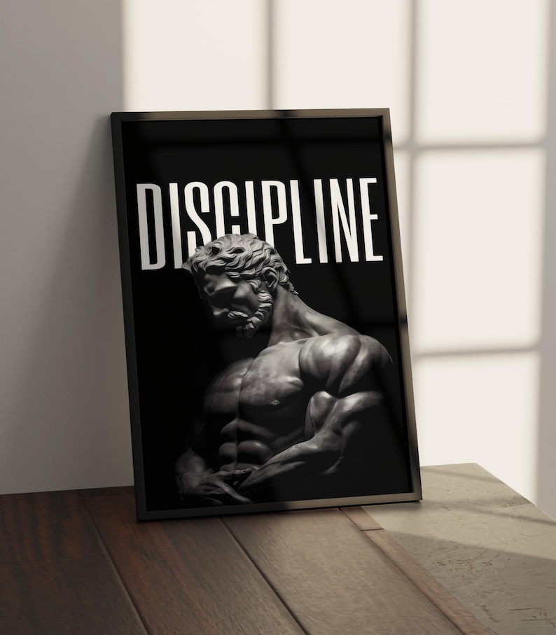 May include: Framed black and white print featuring a muscular statue of a man. The word "DISCIPLINE" is printed in large white letters at the top. The print is displayed on a wooden surface.