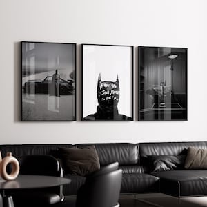 May include: Three framed black and white art prints featuring Batman imagery. One shows Batman near a car, another a Batman mask with text, and the third a Batman figure in a room. The prints are displayed above a black leather sofa.