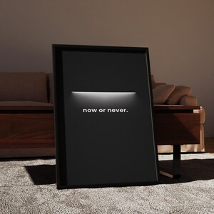 May include: A framed black poster with the words "now or never" in white text. The poster has a horizontal white light bar across the top. The poster is leaning against a wall, with a couch and table in the background.