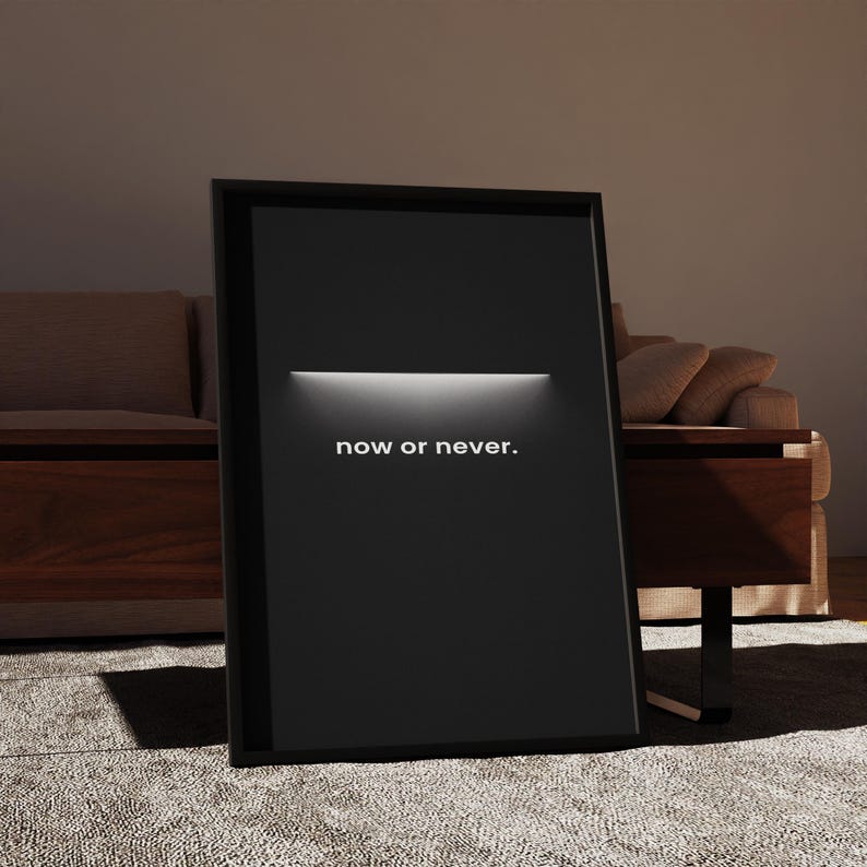 May include: A framed black poster with the words "now or never" in white text. The poster has a horizontal white light bar across the top. The poster is leaning against a wall, with a couch and table in the background.