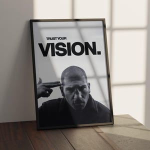 May include: Black and white framed print featuring a man with a serious expression, with the text "TRUST YOUR VISION." The image is leaning against a wall on a wooden surface. The man is pointing his finger to his head.