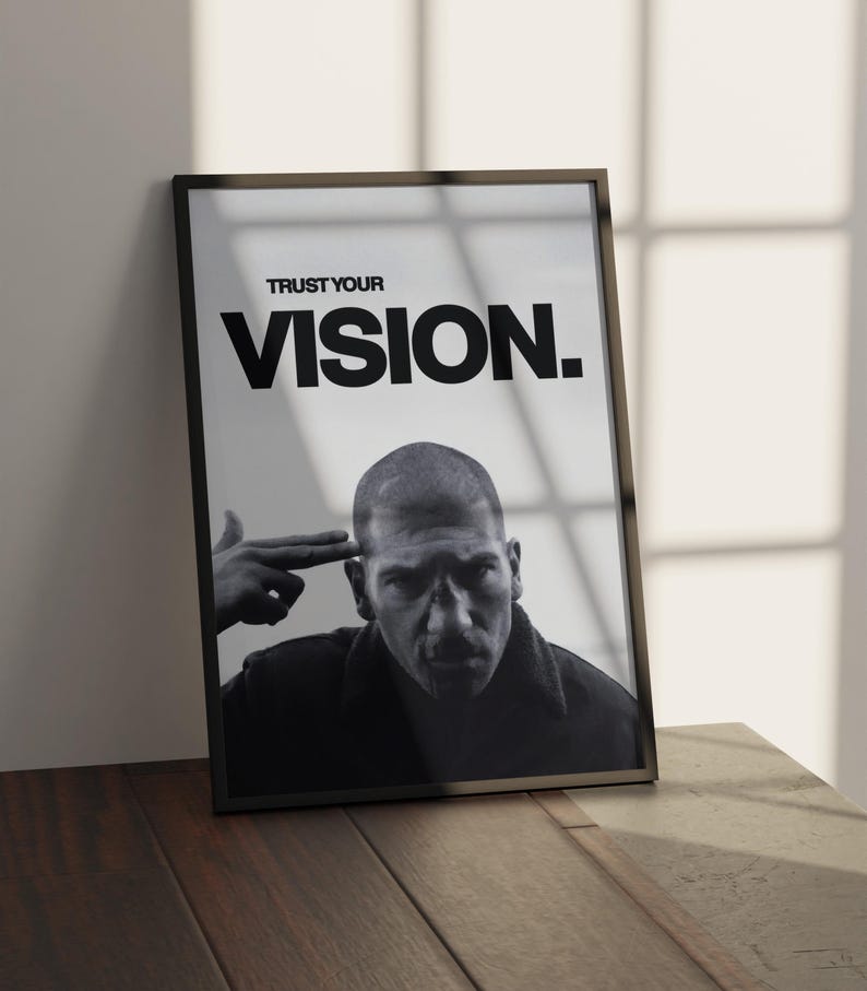 May include: Black and white framed print featuring a man with a serious expression, with the text "TRUST YOUR VISION." The image is leaning against a wall on a wooden surface. The man is pointing his finger to his head.