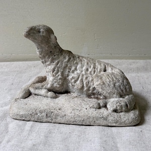 May include: A light gray, textured stone sculpture of a lamb in a resting position. The figure features a rough, weathered surface finish, depicting the animal lying down with its head turned slightly upward, mounted on a rectangular base.