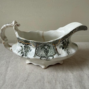 May include: A white ceramic gravy boat featuring a dark green floral transferware pattern with gold trim accents. The vessel has a curved handle, a wide pouring spout, and a scalloped base with four small feet, showing a visible vertical crack.