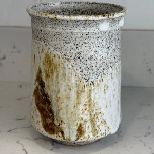 May include: A speckled ceramic vase with a wide opening. The vase has a white base with brown and tan accents. The top of the vase is speckled with black and white dots.