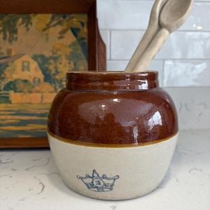 Könnte beinhalten: Ein brauner und cremefarbener Steinguttopf mit einem blauen Kronen-Emblem. Zwei Holzlöffel befinden sich im Inneren des Topfes. Ein gerahmtes Landschaftsbild befindet sich im Hintergrund. Der Topf hat eine glänzende Oberfläche.