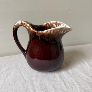 May include: A ceramic pitcher with a glossy dark brown base and a white drip glaze effect around the rim and handle. The pitcher features a curved spout and a rounded body, showcasing a smooth, reflective finish throughout the exterior surface.