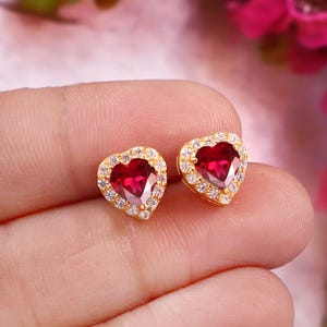 Può includere: Un paio di orecchini a lobo a forma di cuore con una gemma centrale rosso scuro circondata da un alone di piccole pietre chiare e scintillanti, incastonate in una montatura in metallo color oro con chiusura a perno.