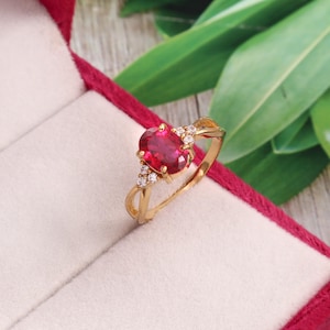 Peut inclure: Une bague en or avec une pierre précieuse rouge foncé de taille ovale fixée par quatre griffes. L'anneau est orné de trois petites pierres claires de taille ronde de chaque côté du sertissage central, créant un design symétrique en métal doré.