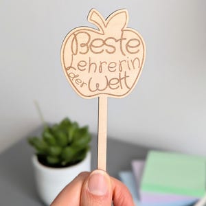Könnte beinhalten: Ein Holzschild in Apfelform mit dem eingravierten Text "Beste Lehrerin der Welt" (Beste Lehrerin der Welt). Das Schild wird von einer Person gehalten. Eine kleine Topfpflanze und Haftnotizen befinden sich im Hintergrund.