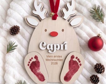 Puede incluir: Un adorno de reno de madera clara con astas blancas, una nariz roja y una cinta de terciopelo rojo. El nombre Capri aparece en letras blancas sobre el texto Mein erstes Weihnachten 2026 y dos huellas de pies de bebé de color rojo.