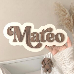 Puede incluir: Un letrero de pared de madera personalizado con el nombre Mateo en letras cursivas de color marr&oacute;n oscuro. Las letras est&aacute;n montadas sobre una base de color crema con forma de nube, creando un efecto de capas con un contraste definido.