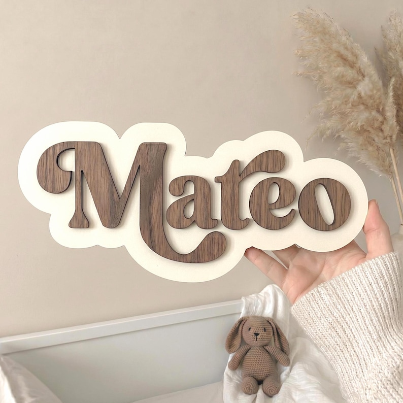 Puede incluir: Un letrero de pared de madera personalizado con el nombre Mateo en letras cursivas de color marr&oacute;n oscuro. Las letras est&aacute;n montadas sobre una base de color crema con forma de nube, creando un efecto de capas con un contraste definido.