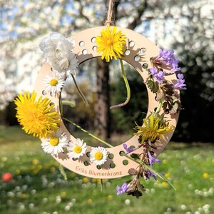 May include: A wooden floral wreath with a variety of flowers, including yellow dandelions, white daisies, and purple wildflowers. The wreath is suspended by a natural twine. The text "Eriks Blumenkranz" is visible on the wooden base.