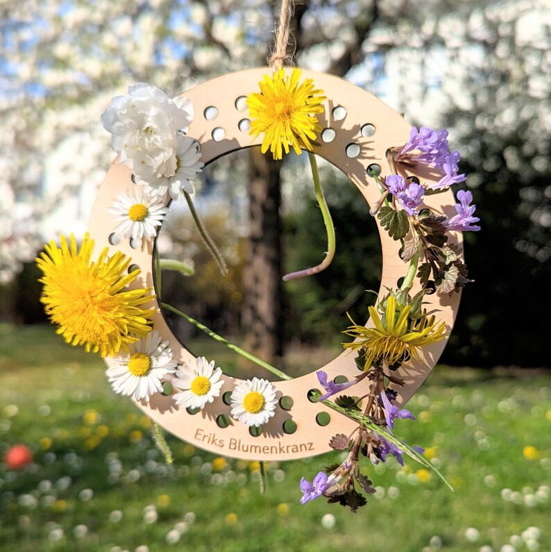 May include: A wooden floral wreath with a variety of flowers, including yellow dandelions, white daisies, and purple wildflowers. The wreath is suspended by a natural twine. The text "Eriks Blumenkranz" is visible on the wooden base.