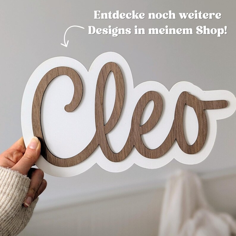 Puede incluir: Un letrero personalizado con el nombre Cleo en una fuente cursiva de madera marr&oacute;n oscuro sobre un fondo blanco. La imagen incluye el texto Entdecke noch weitere Designs in meinem Shop! con una flecha que apunta al letrero.