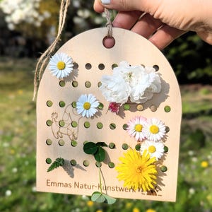 May include: A wooden craft board with small holes, decorated with flowers and greenery. The board features a dog cutout and the text "Emmas Natur-Kunstwerk". A jute string is attached for hanging.