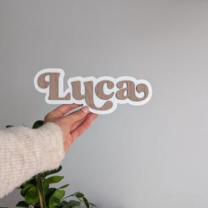 Puede incluir: Un letrero con nombre personalizado que muestra la palabra Luca con un acabado de veta de madera marr&oacute;n, superpuesta sobre un fondo blanco s&oacute;lido. Las letras est&aacute;n conectadas en una fuente redondeada, creando un efecto dimensional con el borde.