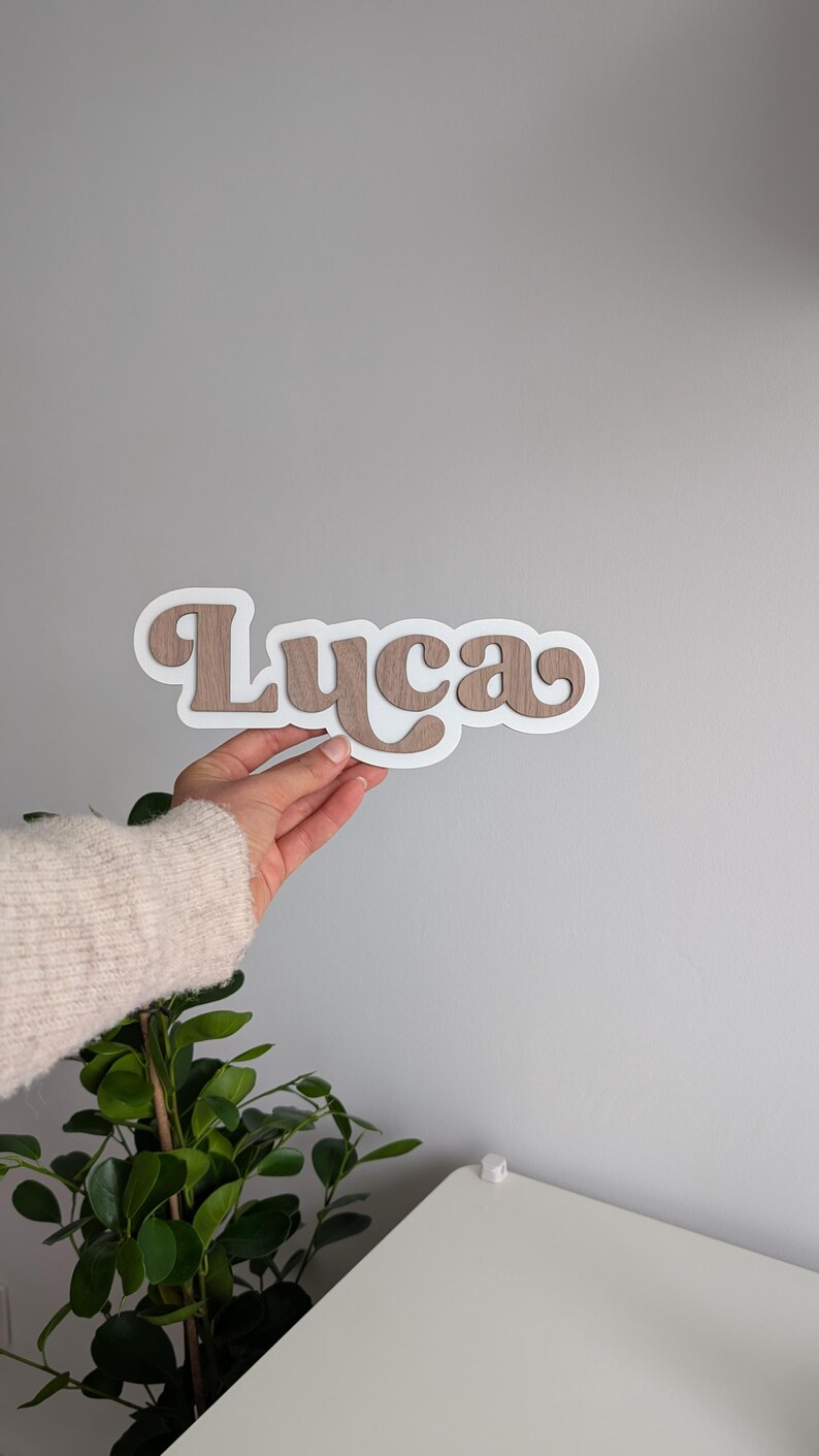 Puede incluir: Un letrero con nombre personalizado que muestra la palabra Luca con un acabado de veta de madera marr&oacute;n, superpuesta sobre un fondo blanco s&oacute;lido. Las letras est&aacute;n conectadas en una fuente redondeada, creando un efecto dimensional con el borde.