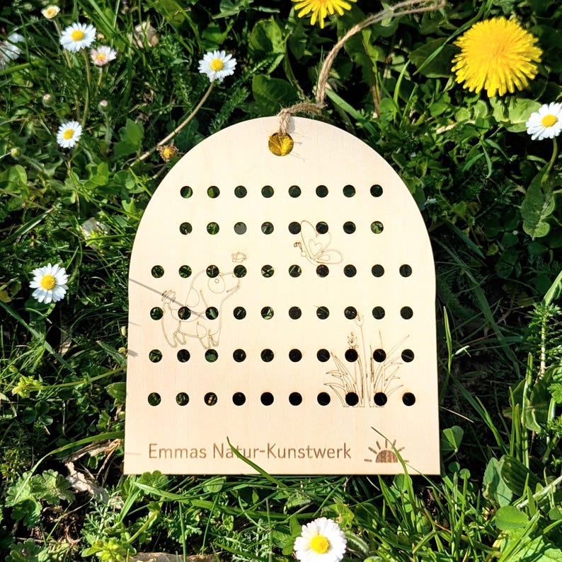 May include: A wooden plaque with a series of evenly spaced holes, featuring etched illustrations of a dog, butterfly, and plants. The plaque has a curved top and a jute string for hanging. The text "Emmas Natur-Kunstwerk" is at the bottom.
