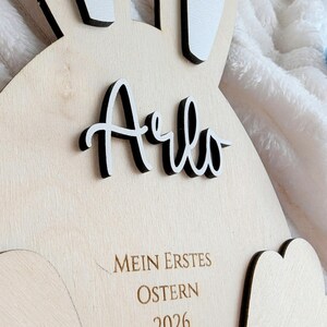 May include: Wooden Easter bunny-shaped decoration with the name "Arlo" in black and white lettering. The text "MEIN ERSTES OSTERN 2026" is engraved below. The bunny has white ears with black trim.