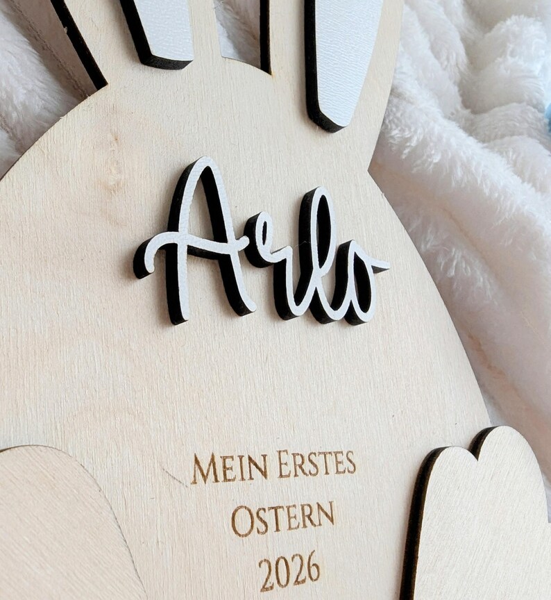 May include: Wooden Easter bunny-shaped decoration with the name "Arlo" in black and white lettering. The text "MEIN ERSTES OSTERN 2026" is engraved below. The bunny has white ears with black trim.