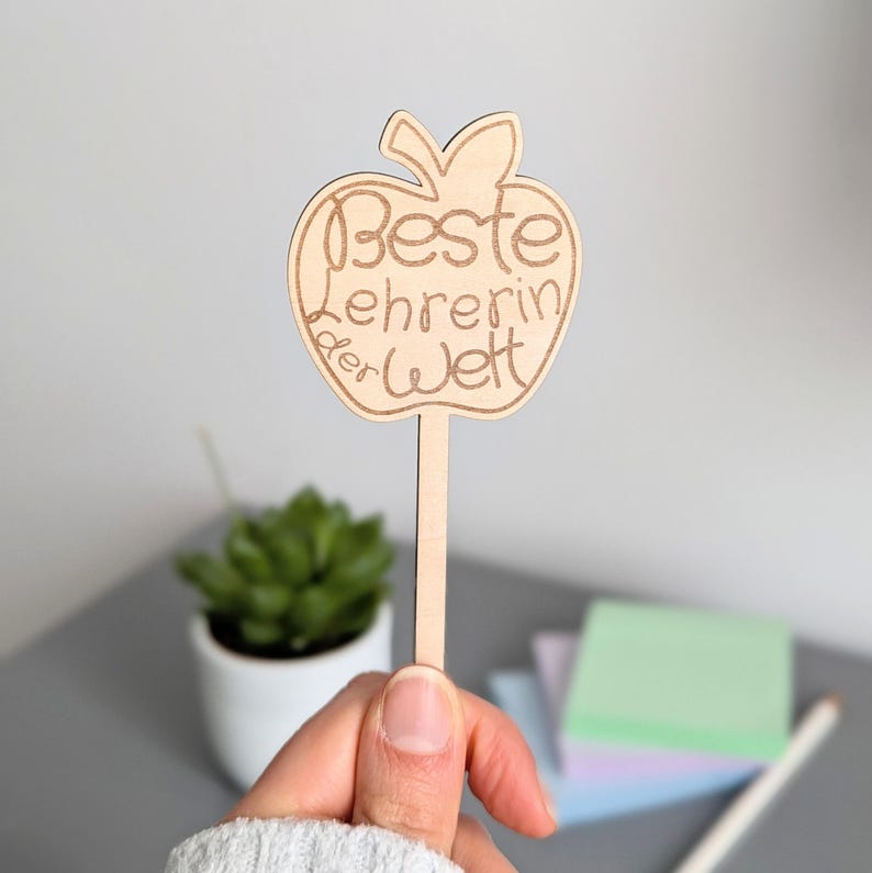 Könnte beinhalten: Ein Holzschild in Apfelform mit dem eingravierten Text "Beste Lehrerin der Welt" (Beste Lehrerin der Welt). Das Schild wird von einer Person gehalten. Eine kleine Topfpflanze und Haftnotizen befinden sich im Hintergrund.