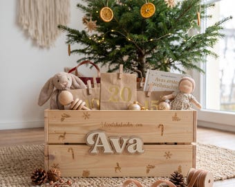 May include: A light-colored wooden crate featuring the engraved text Adventskalender von Ava, decorated with small holiday icons like snowflakes and candy canes. The crate is filled with brown paper bags, a plush bunny, and a small doll.