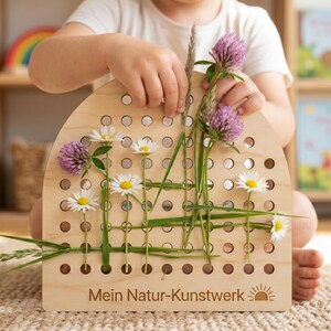 May include: A wooden craft board with a semi-circular top, featuring numerous holes. Daisies, clover, and grass are woven through the holes. The text "Mein Natur-Kunstwerk" is printed on the board. A child's hands are visible, arranging the flowers.