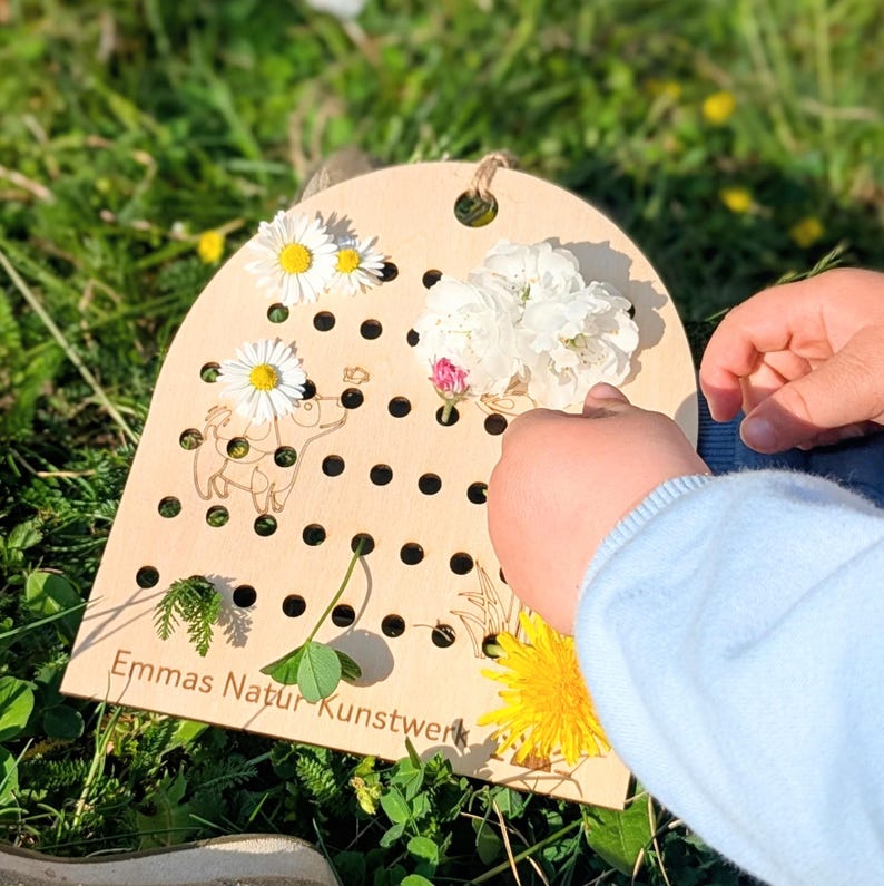 May include: A wooden pegboard with a curved top, featuring a series of small holes and a decorative cutout of an animal. The board is adorned with various flowers and greenery, including daisies and a dandelion. The text "Emmas Natur Kunstwerk" is visible on the board.