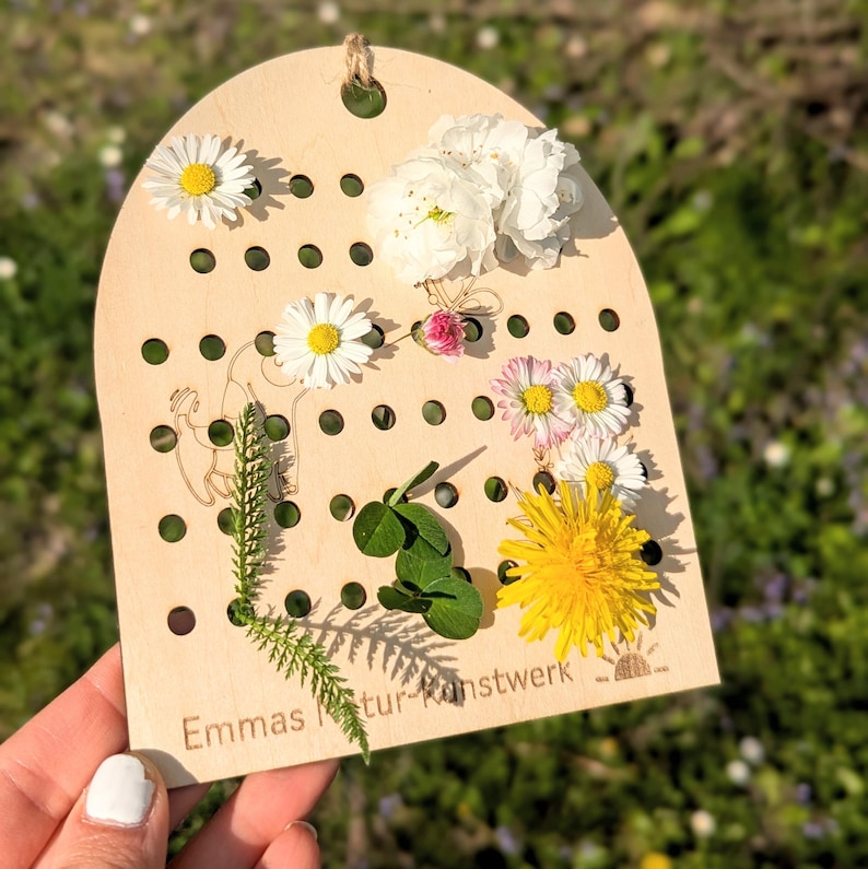 May include: A wooden craft board with small holes, decorated with daisies, clover, and a dandelion. The board has the text "Emmas Natur-Kunstwerk" and a small illustration. A natural, floral arrangement.